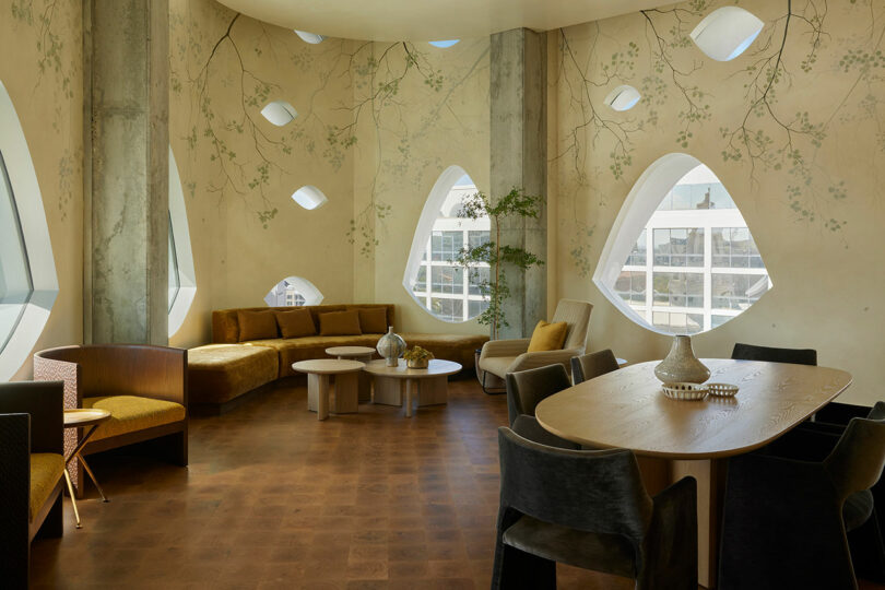 A modern room with unique oval windows, curved seating, and a wooden dining table surrounded by potted plants. The walls feature a subtle floral design, allowing natural light to fill the space beautifully.