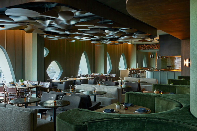 A modern restaurant interior with green curved seating, tables set for dining, large arched windows, and a decorative ceiling. Warm lighting creates an inviting atmosphere.