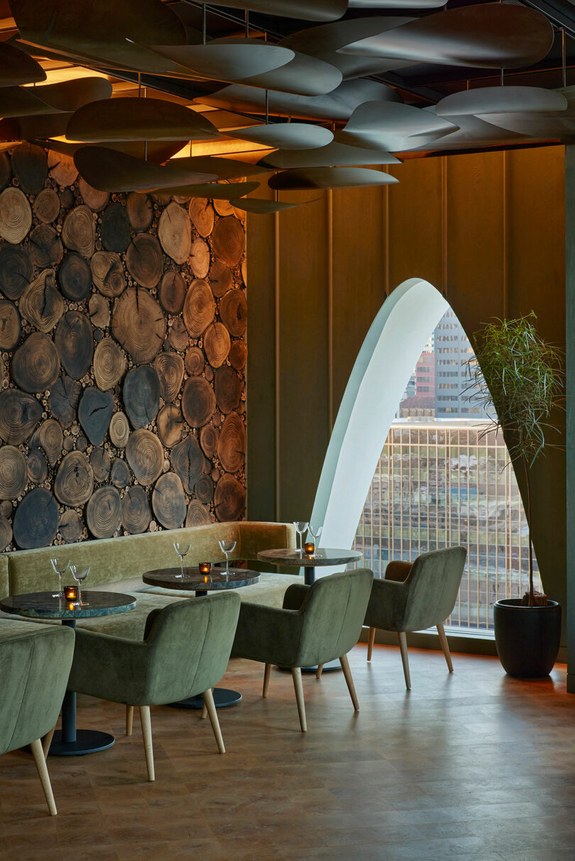 A modern restaurant interior featuring green chairs, round tables, a wall of stacked wood slices, and an arched window that elegantly frames the vibrant urban landscape.