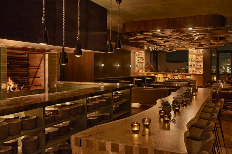 Populus offers a cozy restaurant interior with wooden decor, long bar seating, dim lighting, and an open fire grill. Tables are set with plates and small candlelit decorations, creating an inviting ambiance for every guest.