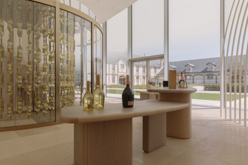 A modern showroom with champagne bottles displayed on wooden tables and a curved glass wall. Large windows reveal a view of buildings and greenery outside.