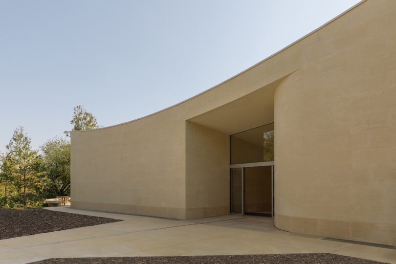 Modern beige building with curved walls and large glass entrance, set against a clear sky and surrounded by sparse trees.