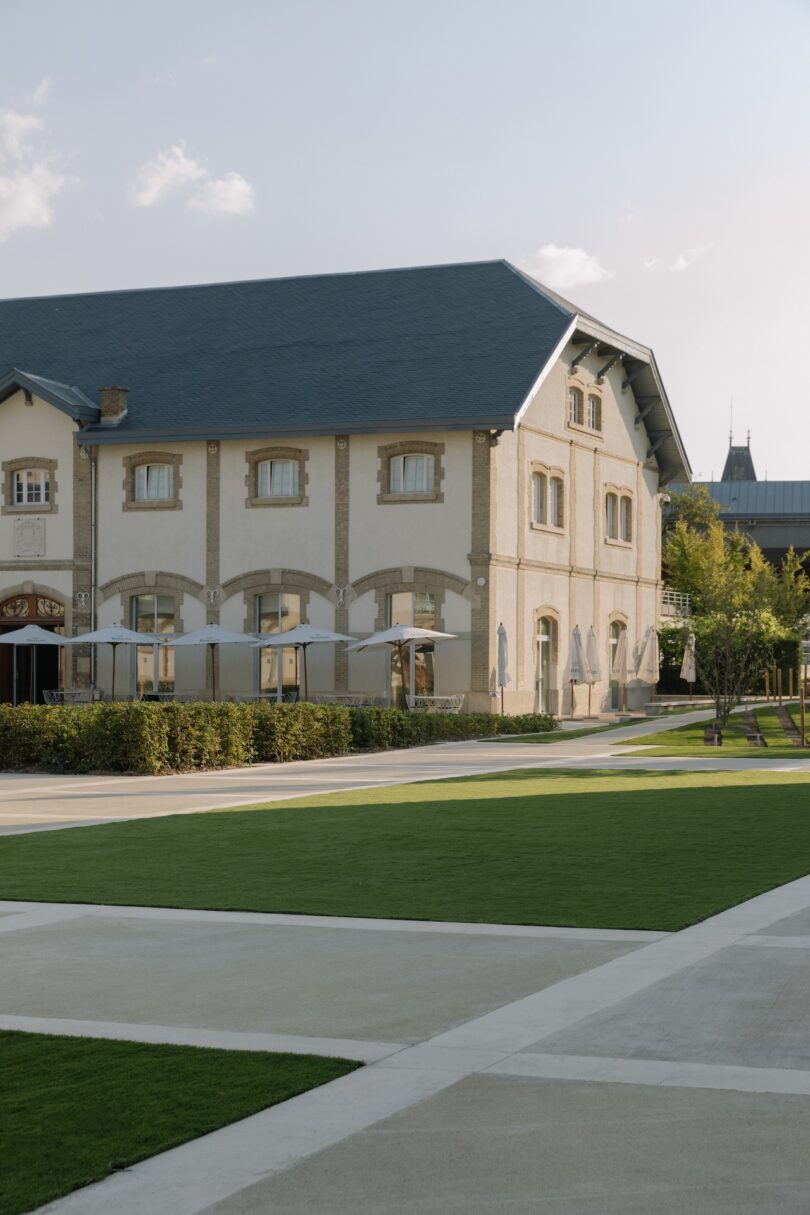 A large beige building with arched windows sits next to a manicured lawn and pathway under a clear sky.