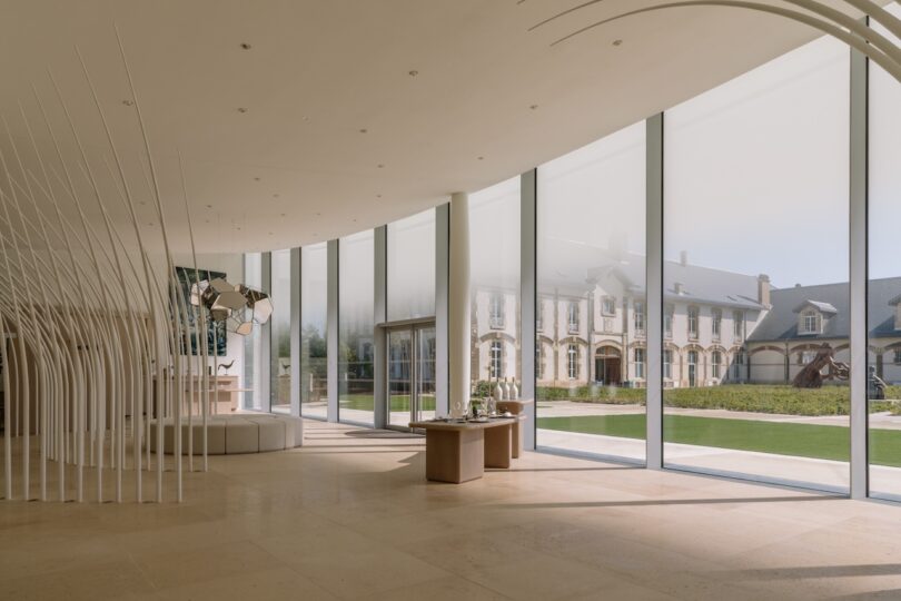 Spacious, modern interior with large curved windows overlooking a historic building. Light-colored flooring and ceiling, minimalistic decor with a table and abstract sculpture.