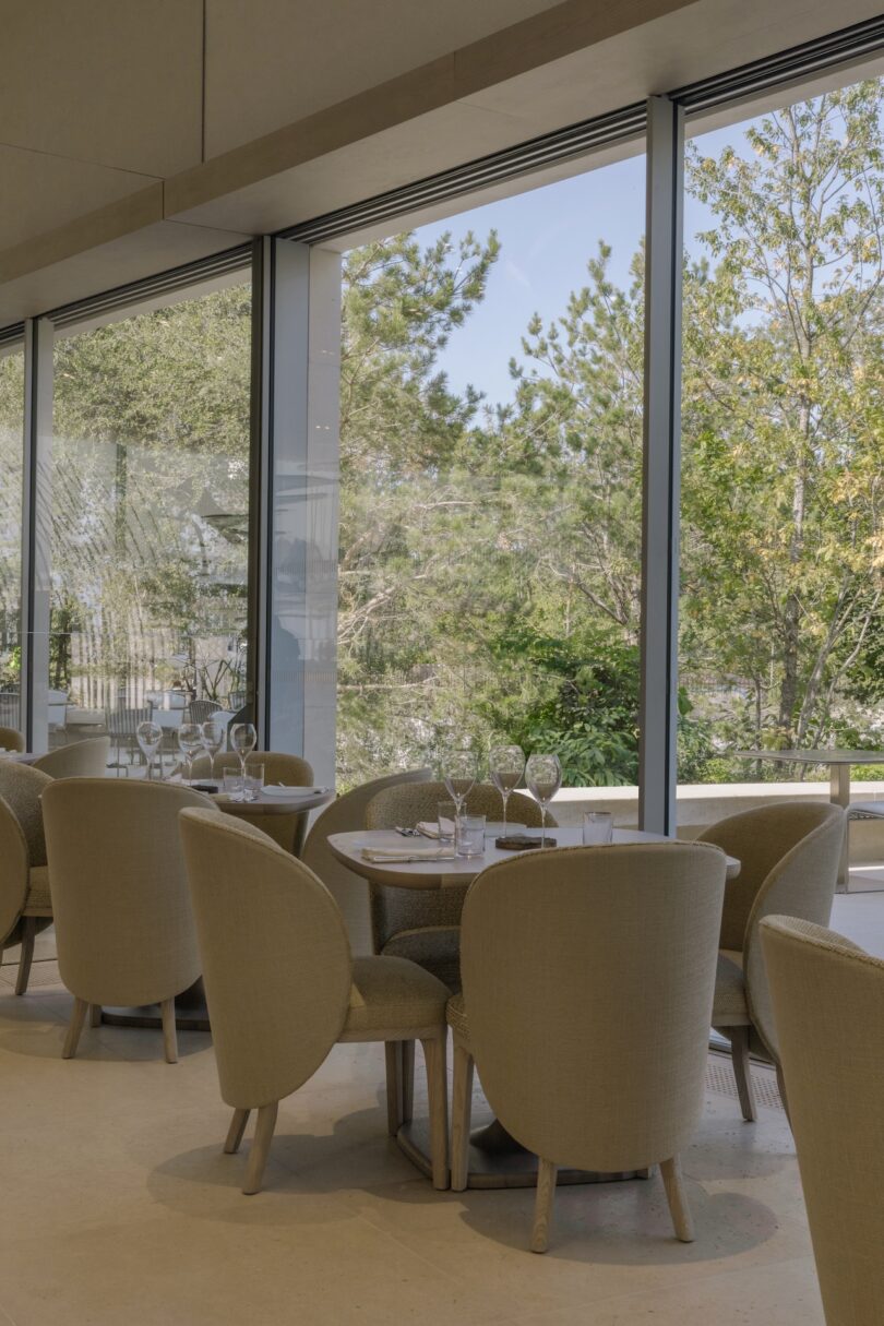 A restaurant interior with beige chairs around circular tables, set by large windows overlooking a garden.