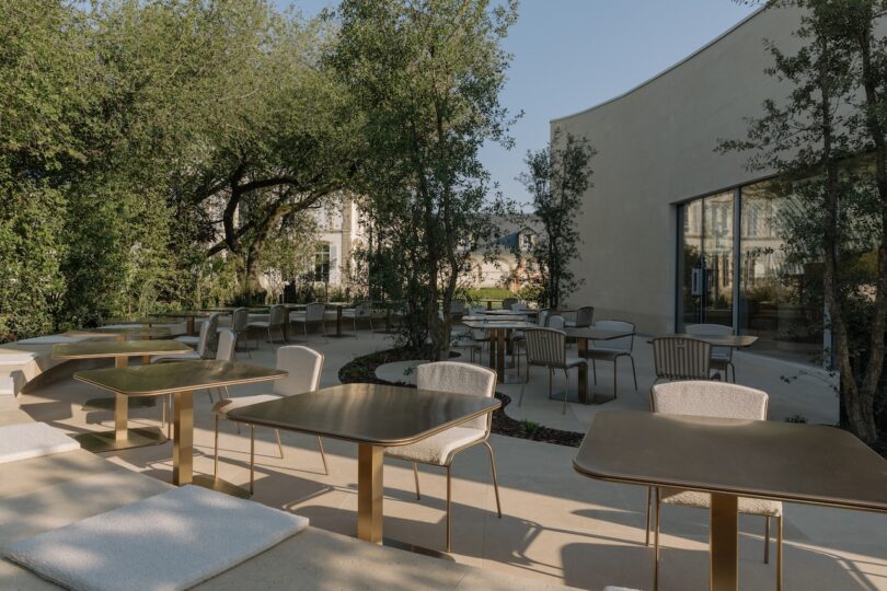 Outdoor patio area with modern tables and chairs surrounded by trees and greenery. Building with large windows in the background.