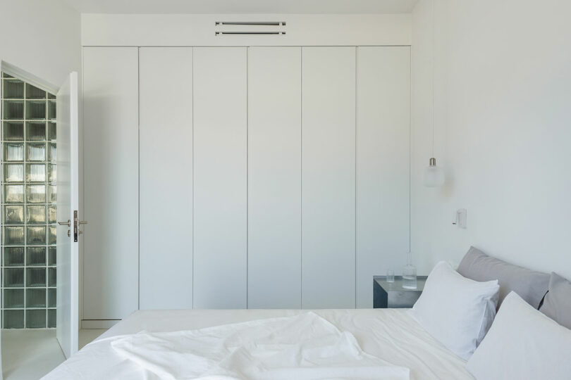 Minimalist white bedroom with a bed, pillows, and a bedside table. Closed wardrobe doors and an open door with glass block walls are visible.