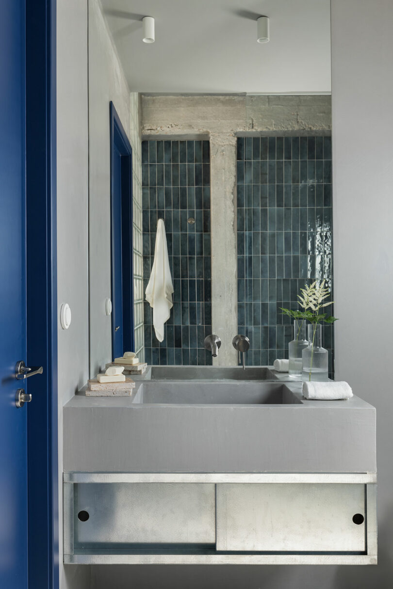 Modern bathroom with a gray concrete sink, wall-mounted faucets, and a large mirror. Blue tiles and a white towel hang in the background. A single flower in a vase is on the counter.