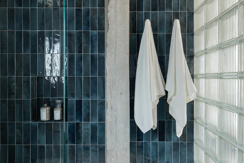 Two white towels hang on a wall next to dark teal tiles and glass blocks. Nearby, two bottles are placed on a shelf.