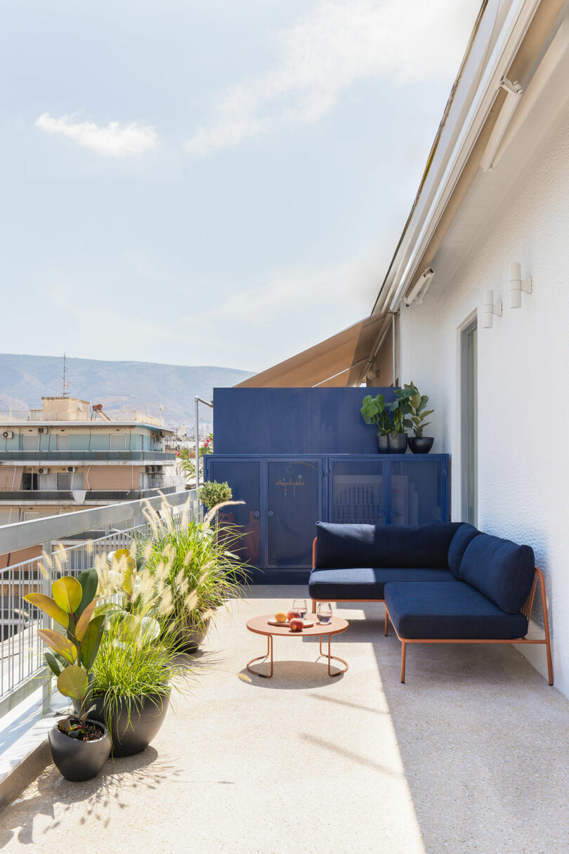 Modern balcony with a blue sectional sofa, round wooden table holding mugs, potted plants, and a view of city buildings and mountains in the background.