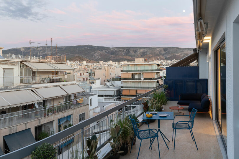 Balcony with a table, chairs, and potted plants overlooking a cityscape with buildings and distant hills under a cloudy sky.