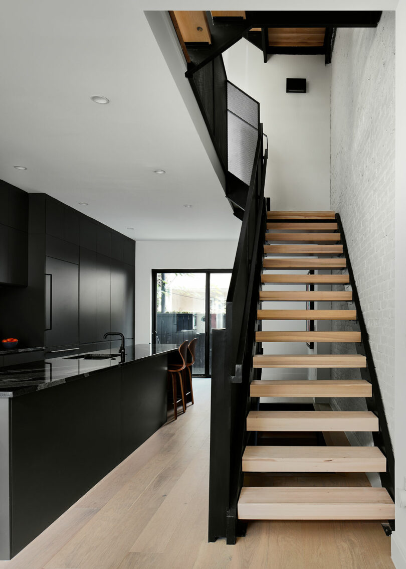 A modern interior featuring a wooden staircase with black metal railings beside a sleek kitchen with dark cabinetry and a large island counter. The space is illuminated by natural light from the window.