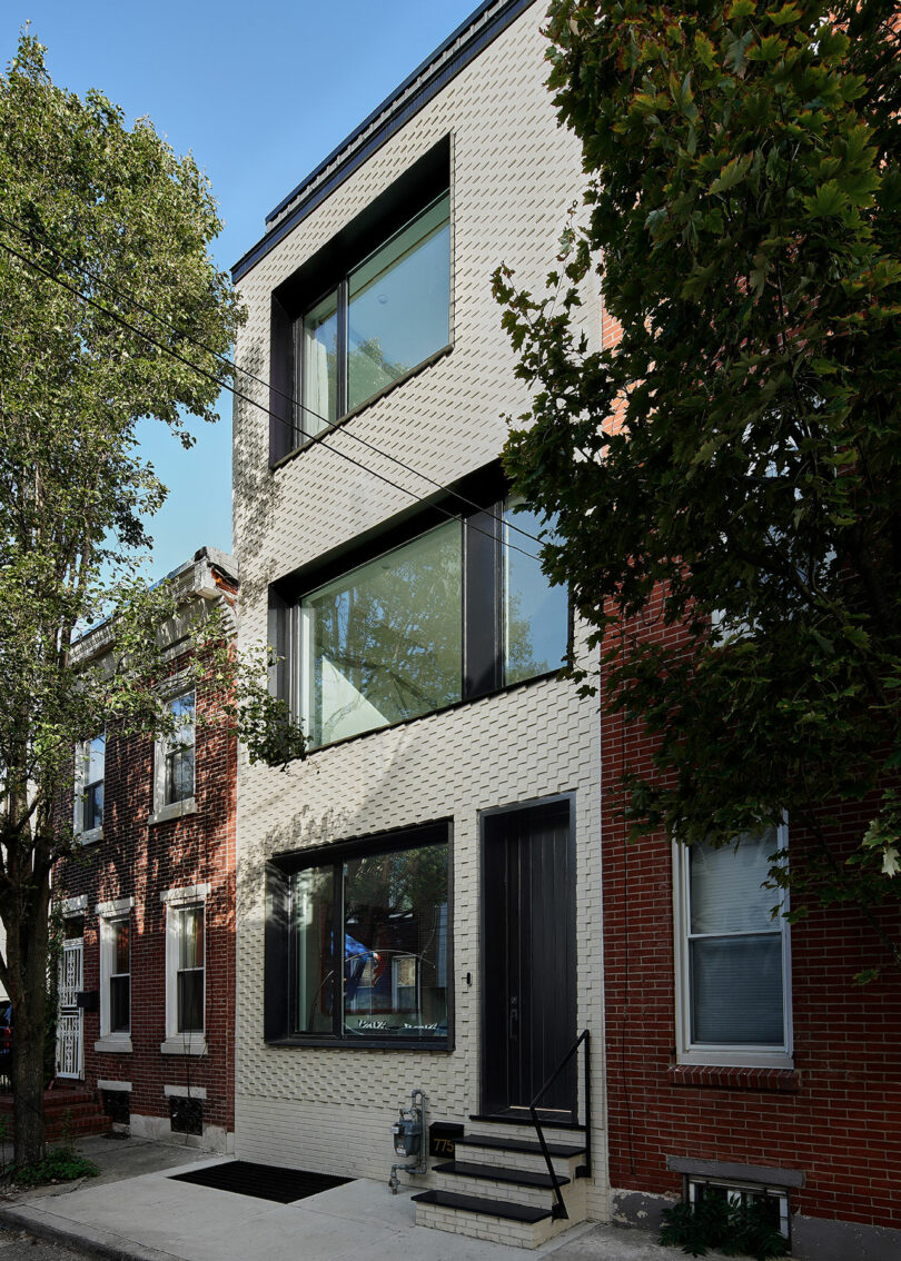 A modern three-story brick townhouse with large windows and a black front door, flanked by older brick houses, and trees lining the street.