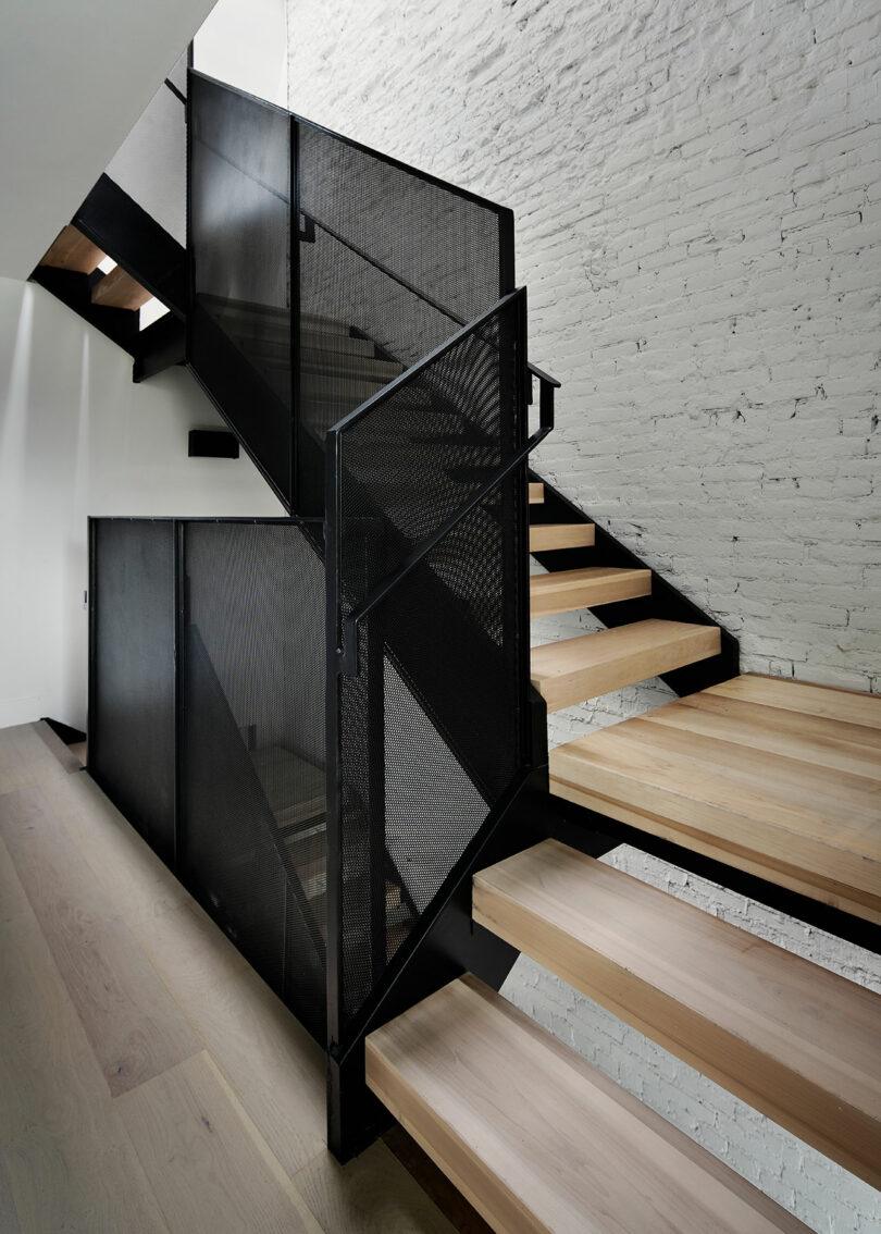 A modern staircase featuring black metal railings and light wooden steps against a white brick wall.