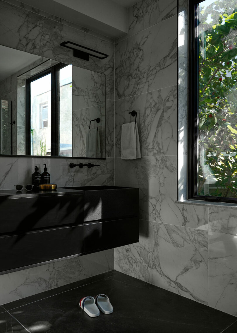 A modern bathroom with marble walls, a black vanity, a large mirror, a window showing greenery outside, and a pair of slippers on the dark tile floor.