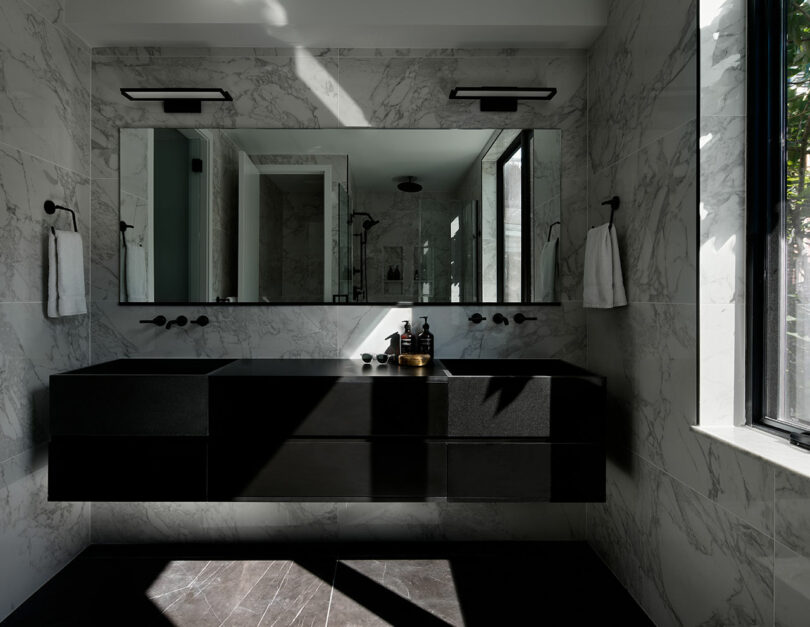 A modern bathroom with marble walls and black vanity featuring double sinks, large mirror, and wall-mounted faucets. Two towels hang on the walls, and natural light filters in through a large window.