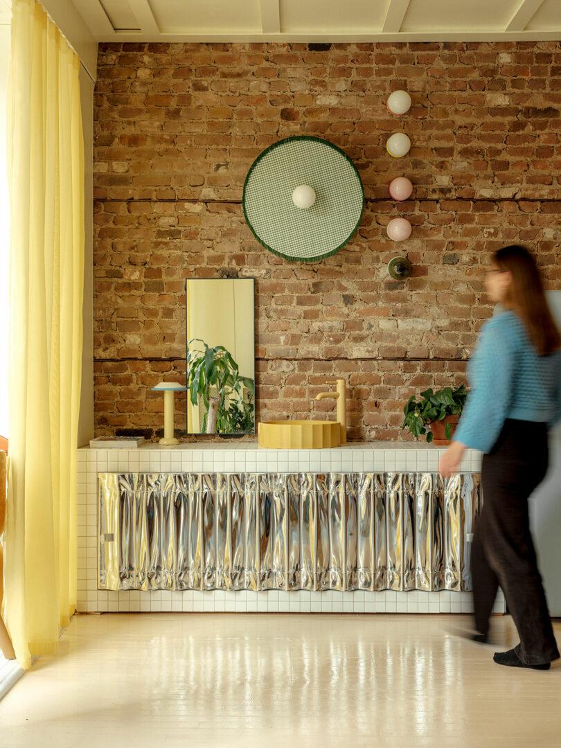 A person in a blue sweater walks past a decorative console with a wavy metallic facade against a brick wall, adorned with round art pieces and a small plant.