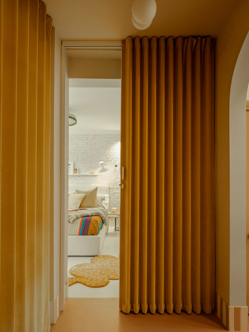 A room with mustard yellow curtains leading to a cozy bedroom featuring a white brick wall, a bed with colorful blankets, and a round rug.