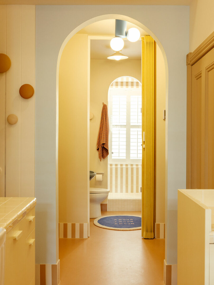 A brightly lit, pastel-toned bathroom with an arched doorway, decorative lights, yellow folding door, and a blue rug on the floor.