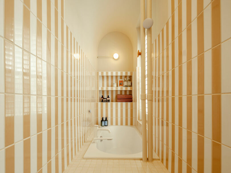 A narrow bathroom with a white bathtub, beige and white striped tiles, built-in shelves with toiletries, a towel, and a round wall light above.