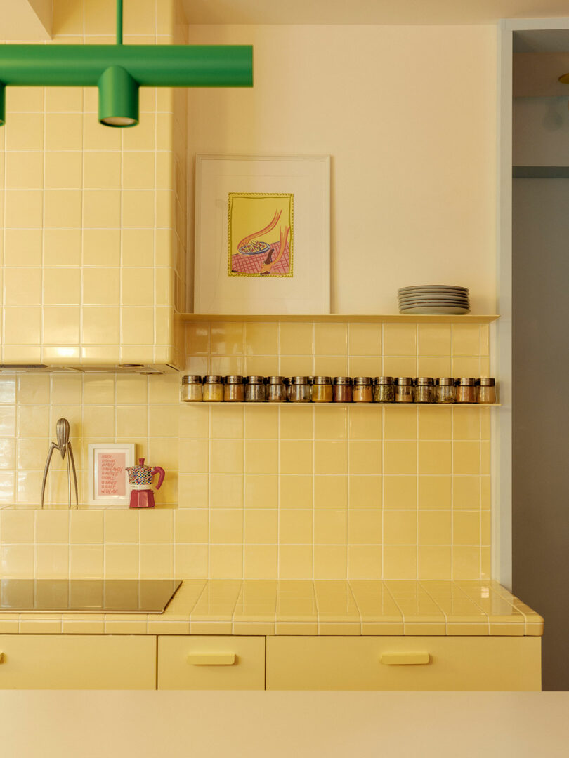 Yellow tiled kitchen with a green lamp, spice jars on a shelf, a framed print on the wall, and kitchenware on the counter.