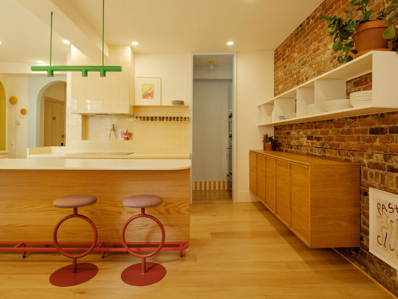 Modern kitchen with a wooden island, two circular stools, brick accent wall, open shelving with dishes, and green pendant lighting.