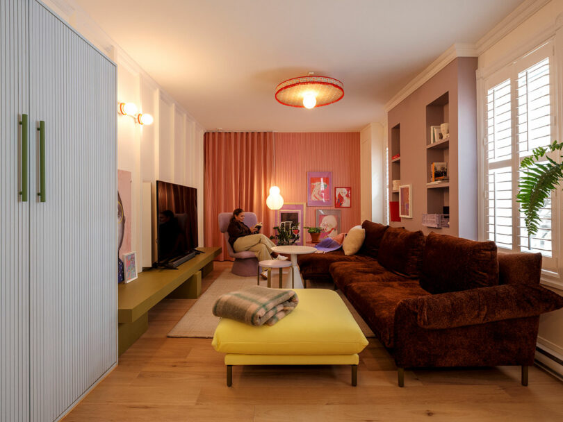 A cozy living room with a brown sofa, yellow ottoman, and a TV. A person sits at a pink table, and colorful decor, including wall art and plants, adds vibrancy to the space.