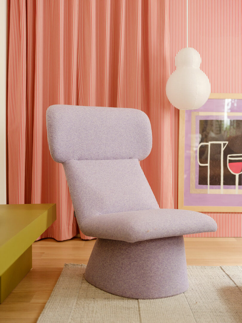 A modern, lavender upholstered chair sits on a beige rug in front of pink striped curtains. A geometric framed art piece and a white spherical lamp are in the background.