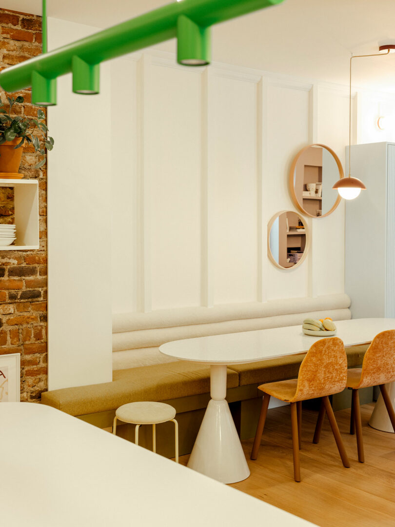 A modern dining area with a white oval table, orange chairs, and a beige bench against a brick and white paneled wall. Green light fixture hangs above. Round mirrors decorate the wall.