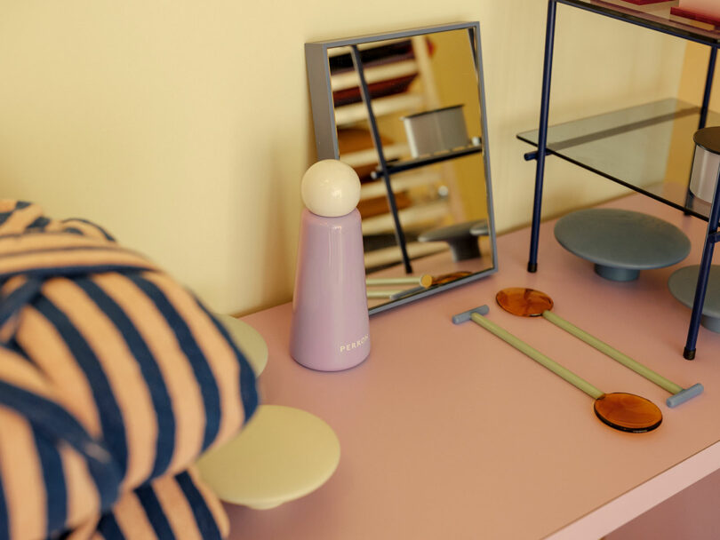 A tabletop with a striped blanket, a small mirror, a pink bottle, and round decorative items on a pink surface.