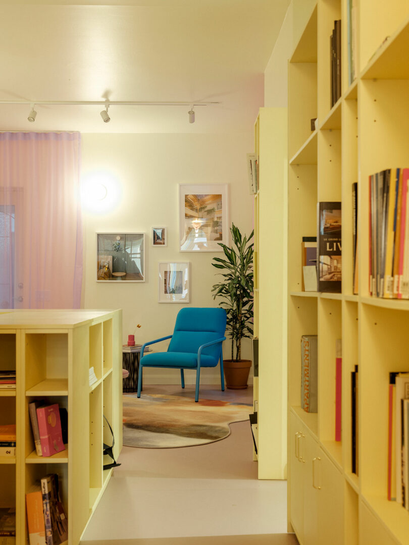 A cozy reading nook with a blue chair, surrounded by yellow bookshelves filled with books. A potted plant and framed art adorn the walls, and soft lighting creates a warm atmosphere.