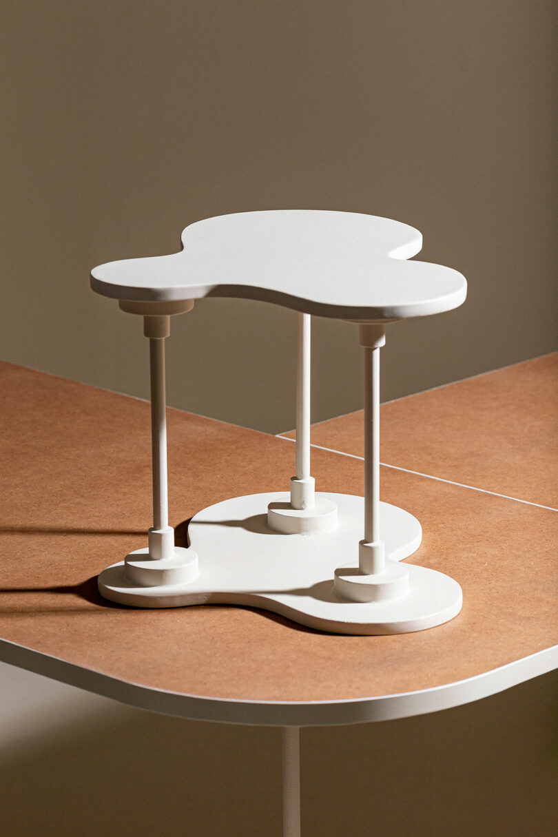 A white, abstract-shaped table with four cylindrical legs stands on a brown surface in front of a beige background