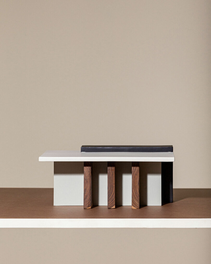 A minimalist architectural model featuring a white rectangular structure with three brown cylindrical supports underneath, set against a beige background