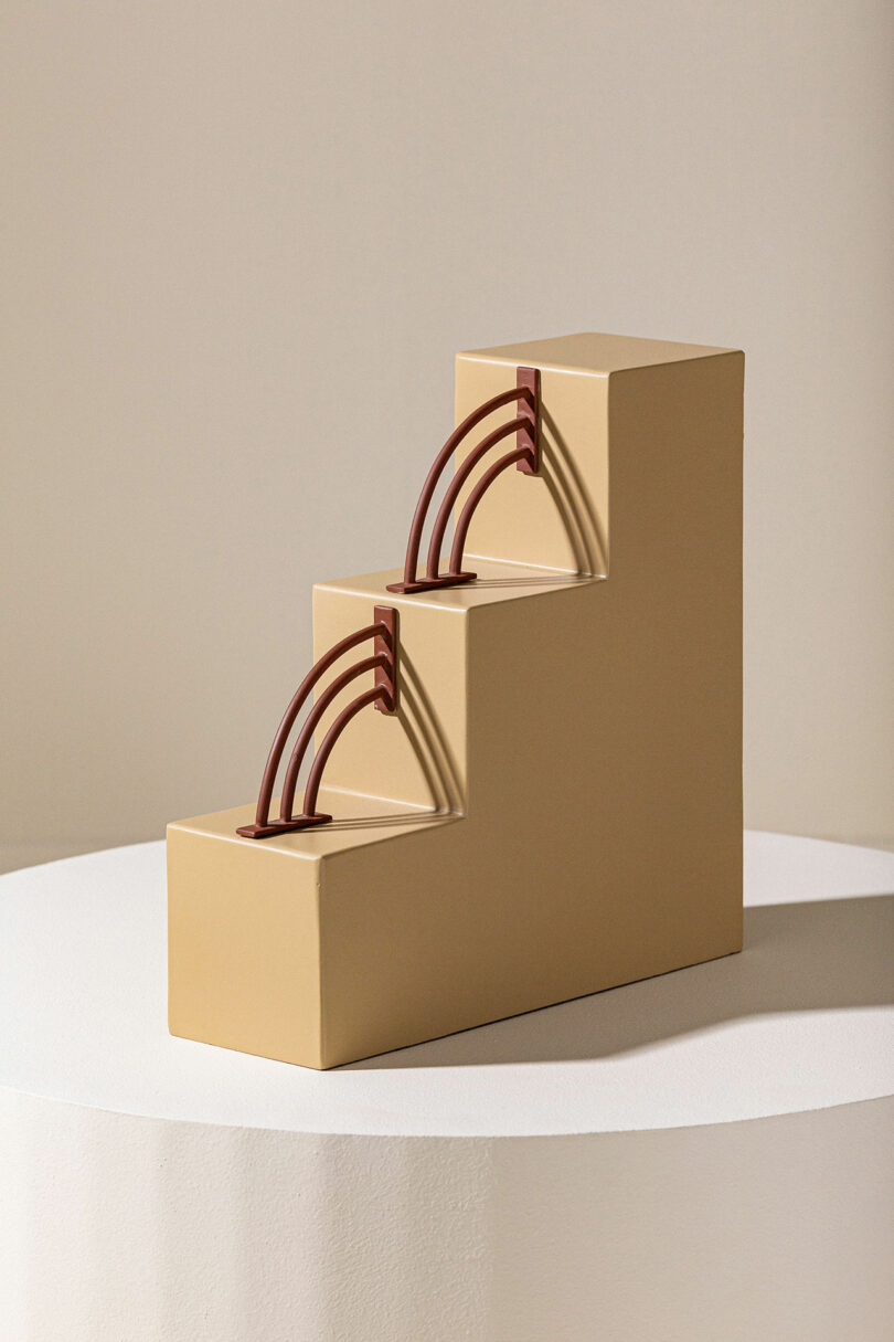 A modern, beige sculpture featuring three ascending blocks, each with an arched, dark red handle, placed on a round white pedestal against a plain background