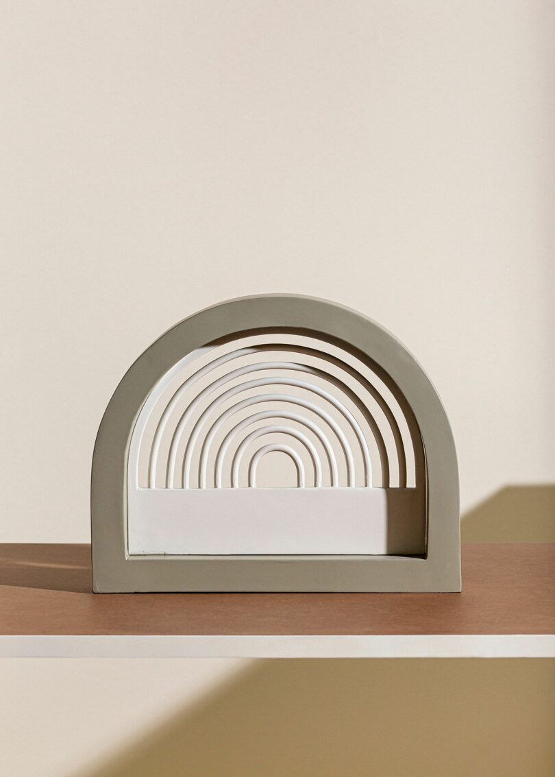 A modern sculpture with concentric arch shapes in beige and white is placed on a table against a neutral background