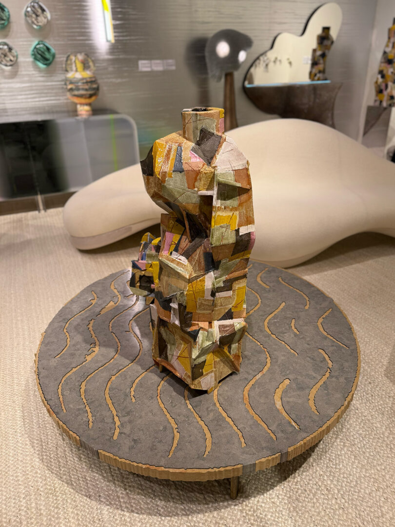 Abstract sculpture made of colorful, irregularly shaped pieces on a circular platform with wavy patterns. A modern, minimalist interior setting with a white couch and various artworks in the background.