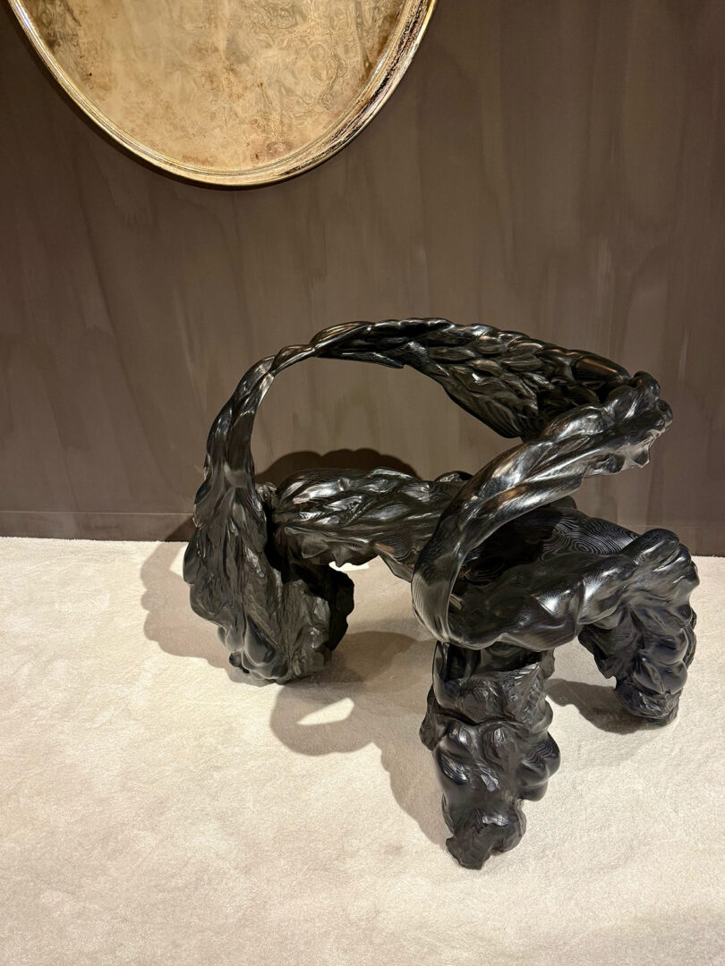 Ornate black sculptural chair with intricate, swirling design sits on a light carpet in front of a dark wall.