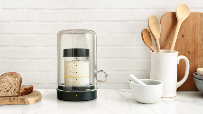 A kitchen counter with a glass-covered fermenting jar, a loaf of bread, wooden utensils in a white holder, and a mortar and pestle.