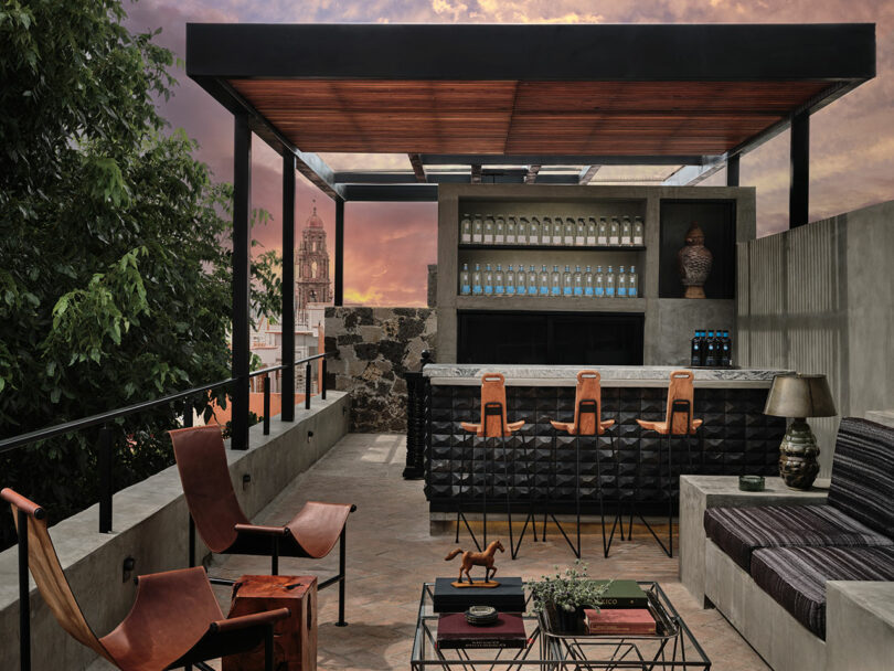 Rooftop bar with modern decor, leather chairs, and a black counter. Bottles are displayed on shelves, and there's a sunset view.