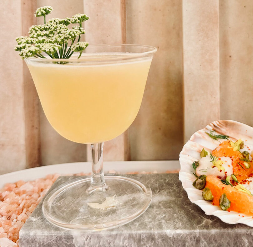 A yellow cocktail in a glass, garnished with small white flowers. Beside it is a shell with orange segments, herbs, and seeds, placed on a stone surface.