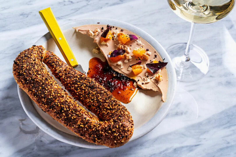 Plate with liver pâté topped with fruit and spices, served with a seeded baguette and a knife. A glass of white wine is to the side.