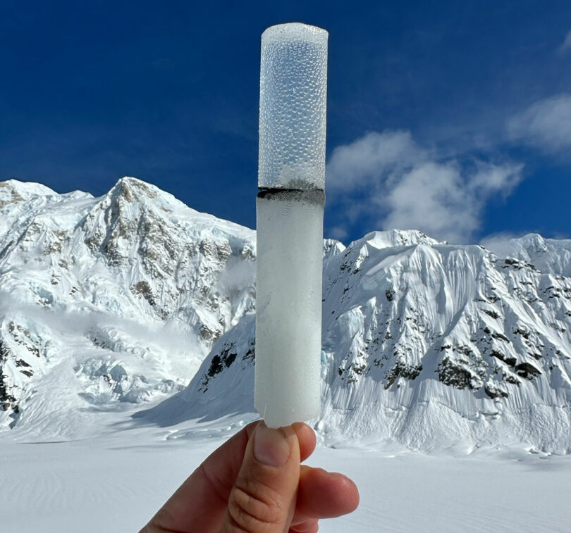 A hand holds an ice core sample with a dark line in the middle, set against a backdrop of snowy mountains under a blue sky.