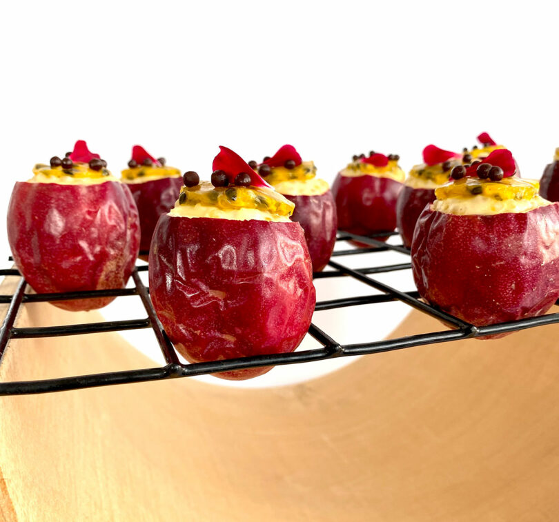 Close-up of red hollowed fruit filled with yellow cream, topped with chocolate pearls and red petal pieces, placed on a wire rack over a wooden surface.