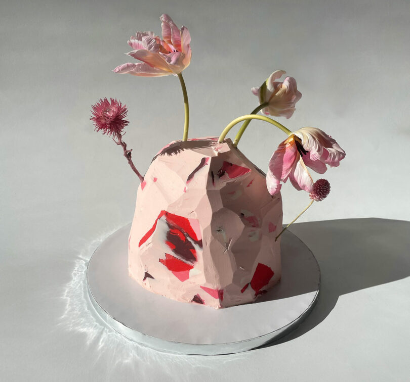 A sculptural pink cake with red and black accents is adorned with drooping pink flowers, displayed on a reflective silver base.