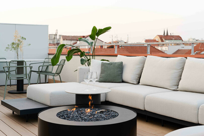 A rooftop terrace with a white sectional sofa, a round fire pit, two glasses on a table, a potted plant, and a dining set in the background. Rooftops and spires are visible beyond the railing.