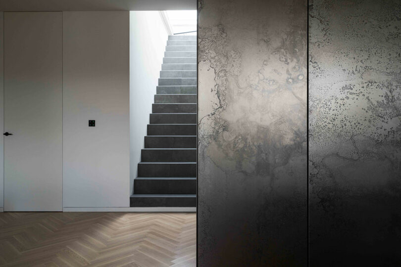 Minimalist interior with a gray staircase, herringbone wooden floor, white walls, and a textured metallic panel on one side.