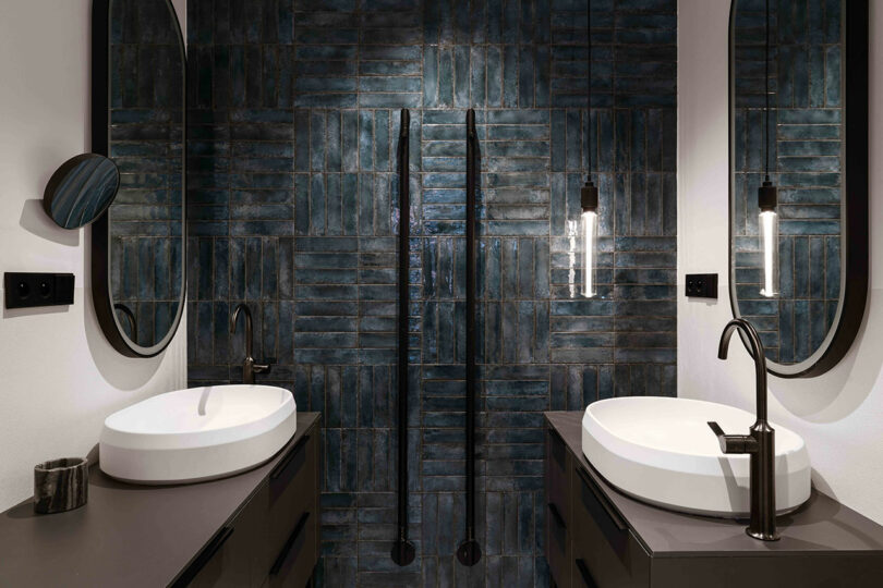 Modern bathroom with two oval mirrors, two sleek sinks, and black fixtures against a dark, textured tile wall. Two pendant lights hang between the mirrors.