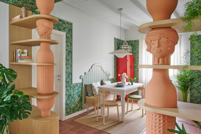 Colorful room with decorative pottery on shelves, green floral wallpaper, and a red door. A dining table has chairs and a plant centerpiece.