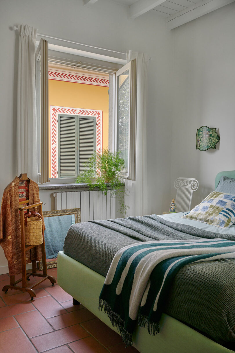 A bedroom with an open window, light-colored walls, a green bed with patterned pillows and blankets, a potted plant on the sill, and a chair and racks with clothes near the bed.