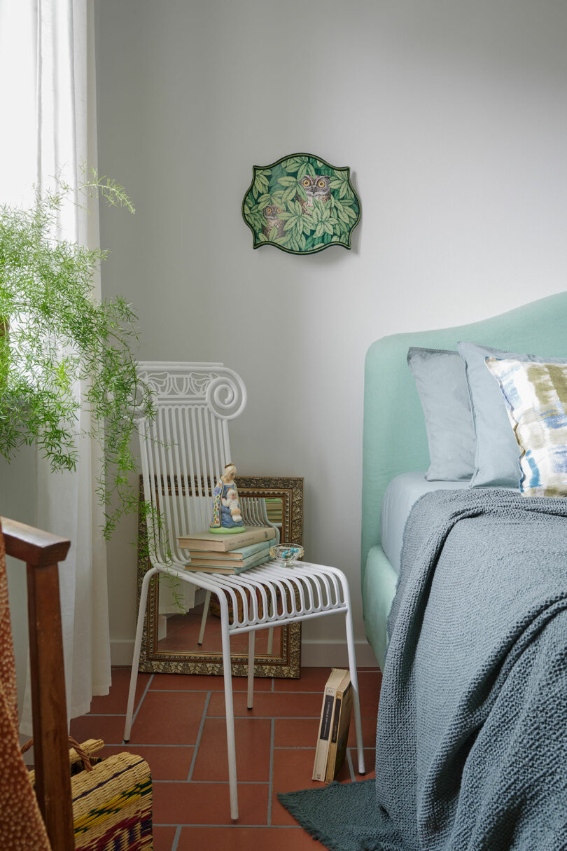 A cozy bedroom with a white chair holding books and decor, next to a mint green bed with blue-gray bedding. A leafy wall art piece hangs above, and a potted plant is nearby.